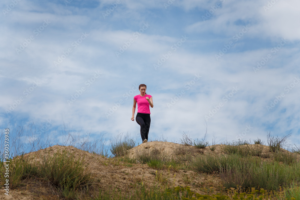 Young sporty woman in sportswear running up hill on cloudy sky background. Fitness, healthy way of life, wellbeing, freedom, mental health, triumph, winning concept.