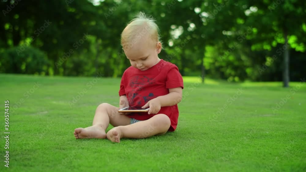 Adorable toddler holding cellphone in hands. Cheerful boy sitting on green grass Adorable toddler holding cellphone in hands. Cheerful boy sitting on green grass