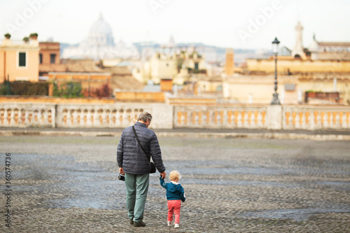 Wall Mural Elderly man holding his small granddaughter hand and amateur video camera walking across old part of Rome