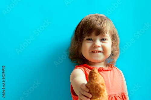 A little cheerful girl in pink dress with curly hair and blue eyes smiles, looks at the camera and holds a croissant in her outstretched hand. The child stands against the blue background.