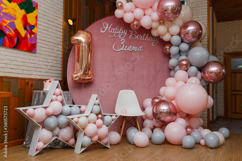 Beautiful festive decorations, pink and grey balloons arch, wooden stars, white chair and number one balloon on wooden round background. Little 1 year old girl birthday party photo zone.