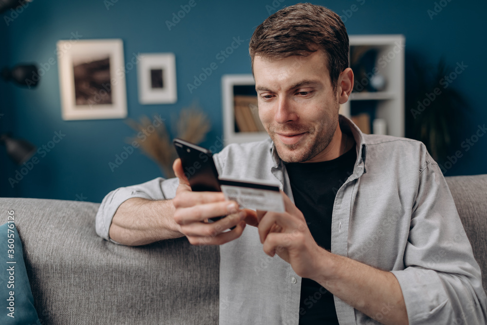 Happy bearded guy doing online shopping while sitting on couch at home. Young hipster in casual outfit using smartphone and credit card for purchase. Happy bearded guy doing online shopping while sitting on couch at home. Young hipster in casual outfit using smartphone and credit card for purchase.