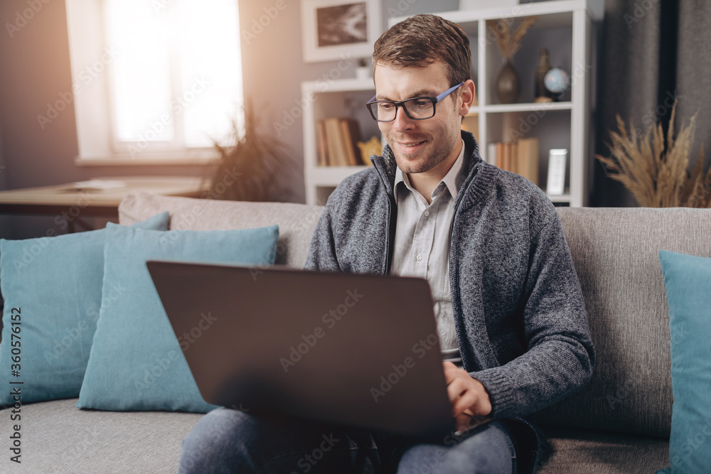 Smiling bearded guy in casual outfit working on laptop while sitting on grey couch. Young male using portable computer for remote work at home. Smiling bearded guy in casual outfit working on laptop while sitting on grey couch. Young male using portable computer for remote work at home.