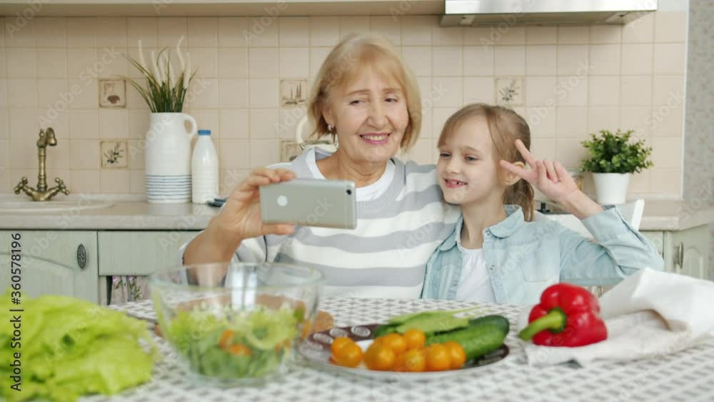 Grandmother and granddaughter are taking selfie in kitchen with smartphone camera showing hand gestures posing smiling. People and devices concept. Grandmother and granddaughter are taking selfie in kitchen with smartphone camera showing hand gestures posing smiling. People and devices concept.