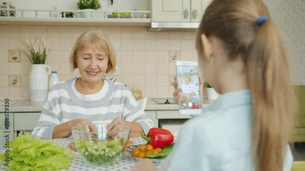 Cheerful elderly woman is posing for smartphone camera and cooking salad while child is taking picture with modern device. Family, photograph and people concept. Cheerful elderly woman is posing for smartphone camera and cooking salad while child is taking picture with modern device. Family, photograph and people concept.