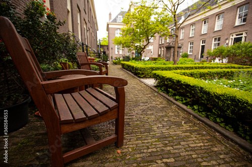 Close up image of a red wooden chairs on the side of a courtyard garden in a senior housing community. In the background there is  a well maintained garden, brick buildings and cobblestone ground.