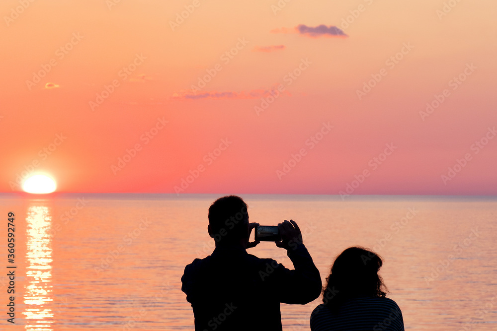 A man photographs an orange sunset on a smartphone. Silhouettes of women and men on a blurred background.