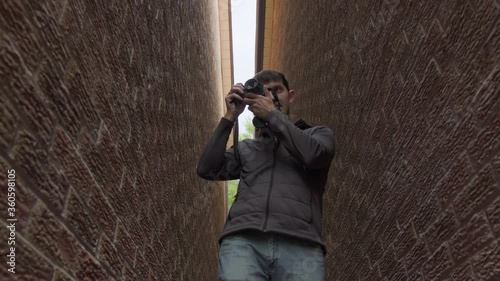 High quality 4k video of a young man who is standing between two red brick walls and holding photo camera in his hands and trying to make a photo or video. Young photographer work in process.