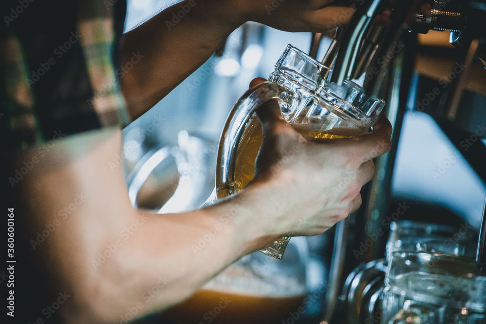 Hand of man pouring tankard of frothy draught beer from stainless steel beer tap in bar or pub into large glass tankard. Bartender at beer tap pouring beer in glass serving in restaurant.