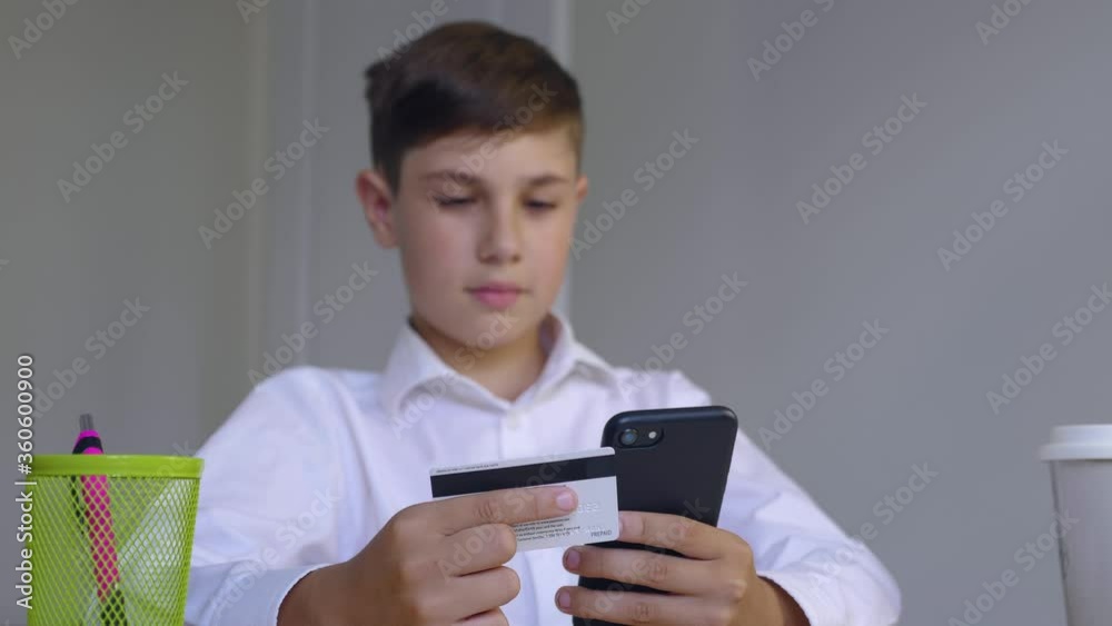 Beautiful boy 10 years old sitting at home makes online payments through the Internet from bank card on smartphone. Child makes purchases in the online store. Online banking with smart phone. Beautiful boy 10 years old sitting at home makes online payments through the Internet from bank card on smartphone. Child makes purchases in the online store. Online banking with smart phone.