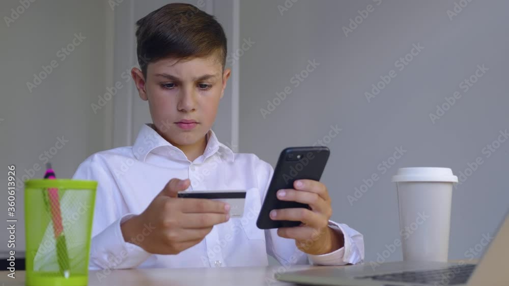 Beautiful boy 10 years old sitting at home makes online payments through the Internet from bank card on smartphone. Child makes purchases in the online store. Online banking with smart phone. Beautiful boy 10 years old sitting at home makes online payments through the Internet from bank card on smartphone. Child makes purchases in the online store. Online banking with smart phone.