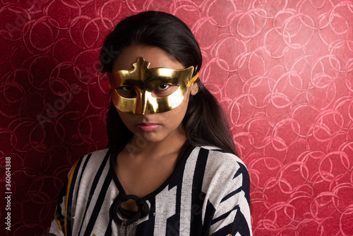 Fashion portrait of an Indian Bengali beautiful and young girl in western dress wearing an eye mask standing in front of a red textured wall. Indian fashion and portrait