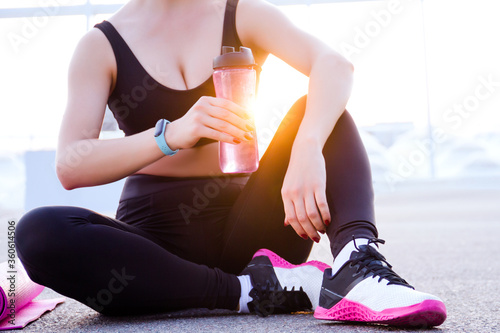 Closeup young athletic woman in black tracksuit is sitting outdoor at sunset, holding pink bottle of water. Girl is relaxing after training, stretching, yoga. Daily routine. Sport lifestyle.