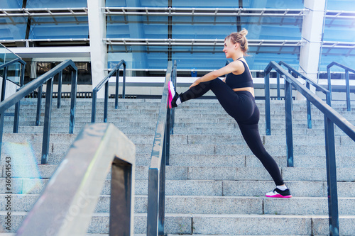 Young athletic woman in black tracksuit uniform is training outdoor on street. Blonde girl is stretching legs muscles on steps of stadium. Daily morning routine. Healthy and sport lifestyle concept.