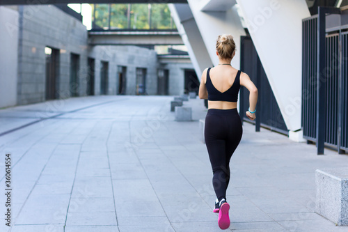 Young athletic woman in black tracksuit is training, doing exercise outdoor at modern city. Blonde girl is jogging, running, preparing for marathon. Daily morning routine. Sport and healthy lifestyle.