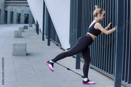 Young athletic woman in black tracksuit is doing stretching exercises for legs muscles with elastic. Girl is training outdoor at modern city. Daily morning routine. Sport and healthy lifestyle.