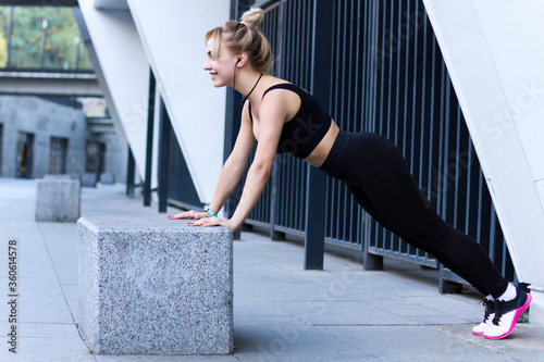 Young athletic woman in black tracksuit is doing gymnastics strength exercises for muscles. Blonde girl is training outdoor at modern city. Daily morning routine. Sport and healthy lifestyle.
