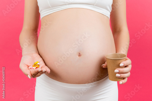 Close up of pregnant woman holding a glass of water in one hand and a pile of pills in another hand at colorful background with copy space. Taking medicine during pregnancy concept