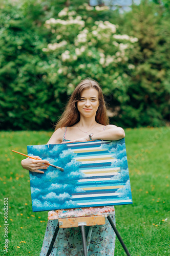 Beautiful girl in blue dress stands near an easel holding a brush. Girl painter artist is painting outdoors. © Yuliia