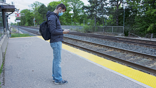 High quality picture of a young man who is waiting for a traing on a railway station. GO train station in Canada. Young handsome guy standing on a station and holding his cellphone wearing  face mask