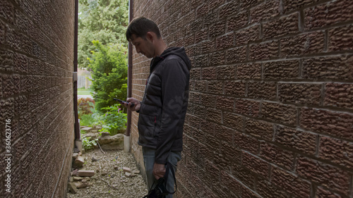 High quality picture of a young man who is texting messages on his cellphone standing in a shade of the red bricks wall. Guy hiding between the walls and holding mobile phone in his hands.