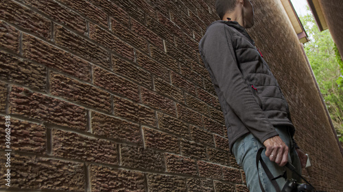 High quality picture of a young man who is standing between two red brick walls and holding photo camera in his hands and trying to make a photo or video. Young photographer work in process.