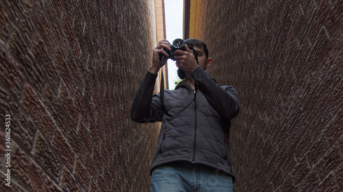 High quality picture of a young man who is standing between two red brick walls and holding photo camera in his hands and trying to make a photo or video. Young photographer work in process.