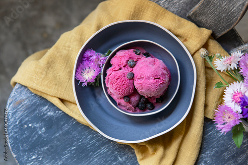 Berry ice cream or blackcurrant sorbet with berries on blue ceramic plate, vintage wooden chair with yellow textile and flowers. Organic raw dessert, healthy food concept. Copy space, top view.