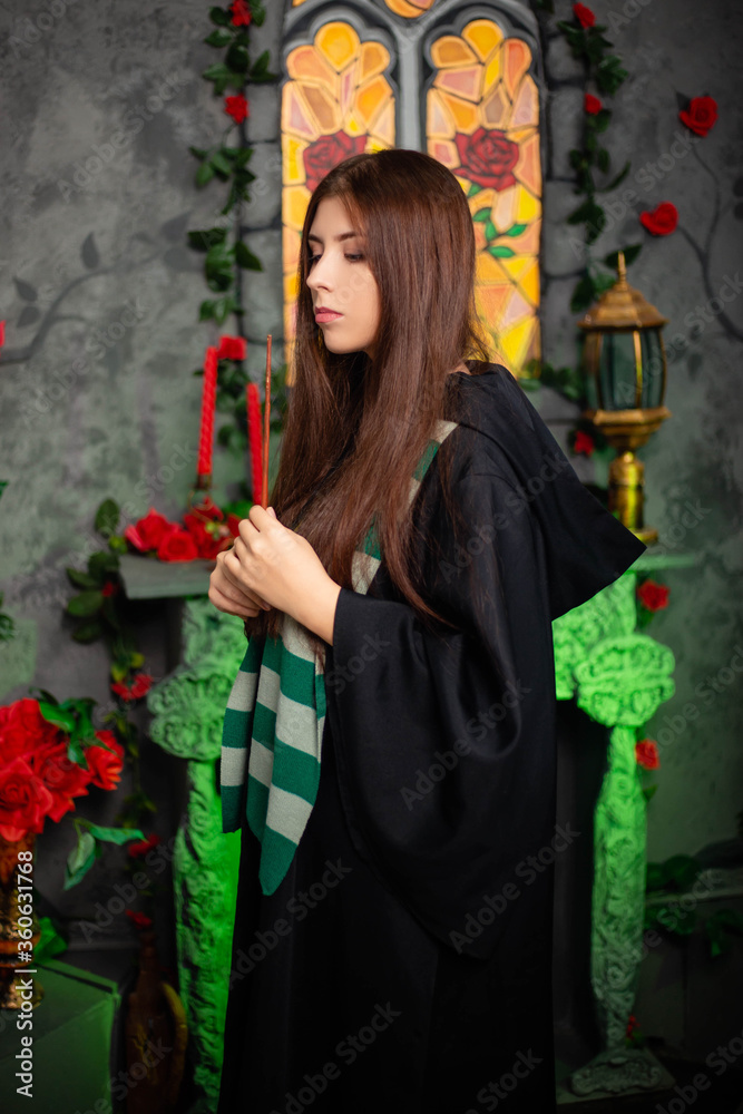 Young woman in a black cloak with a magic wand in her hands, standing against the backdrop of a medieval window. Halloween witch concept, model with clean skin, retouching.