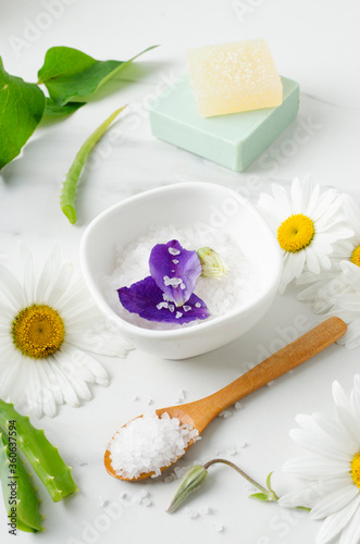 Vertical image.Bowl of sea salt, purple flower petals, chamomiles, wooden spoon, aloe on the marble table