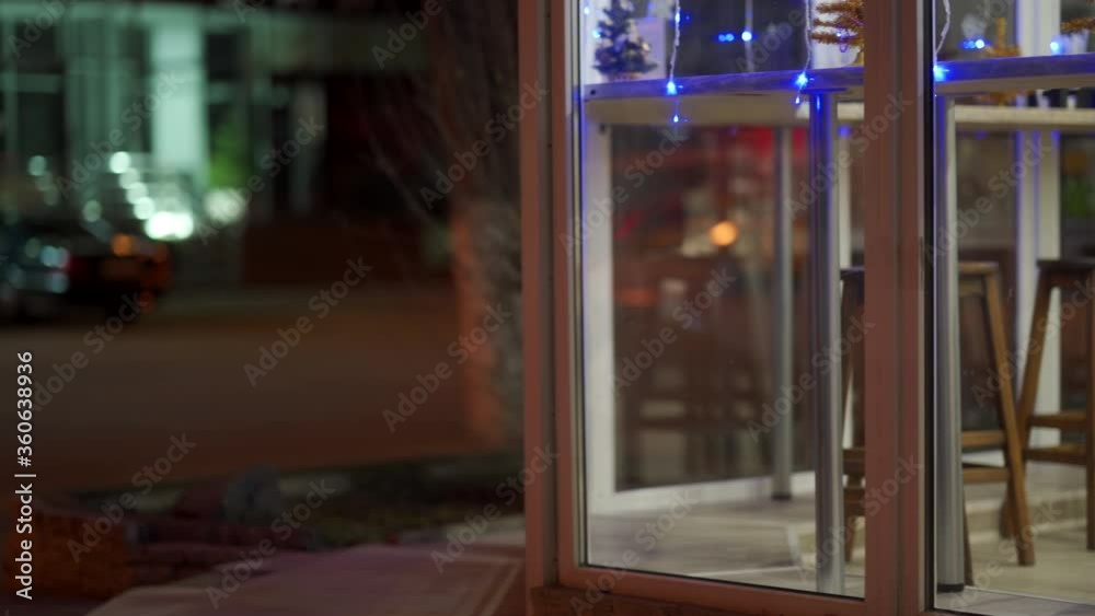 Coffee shop window display with festive lighting at dusk, window of cafe in the evening decorated with Christmas garlands, small Christmas trees on window counter.