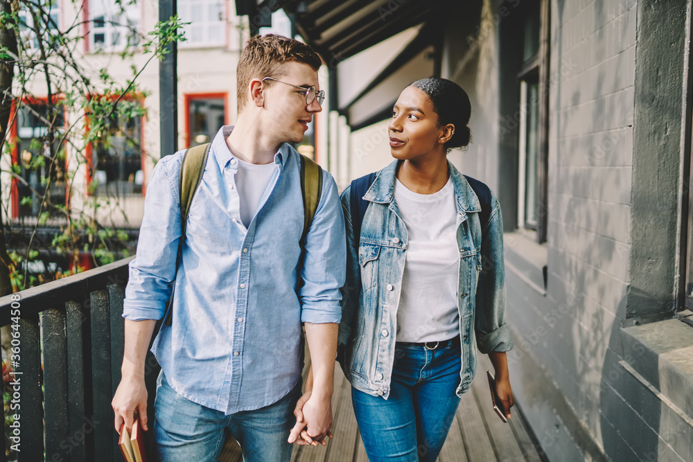 Happy multicultural couple in love dressed in casual wear looking and holding hands each other standing outdoors.Caucasian hipster guy travel together with african american young woman