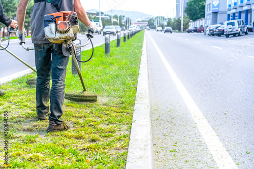 People mow grass with lawn mowers along a roadway.