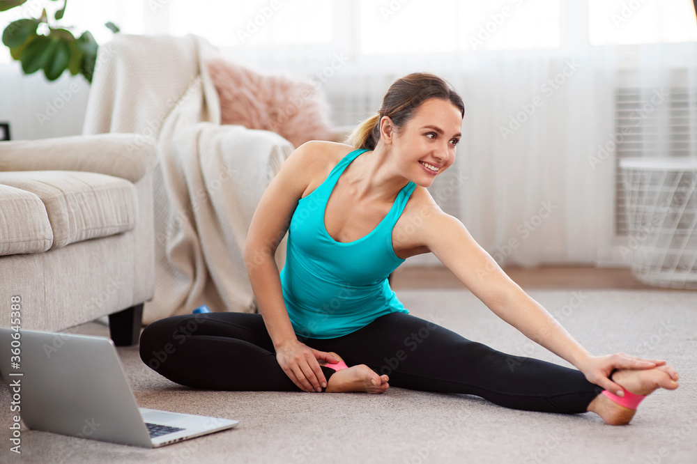 Sporty woman doing fitness routine on floor and watching laptop at home. Online home stretching exercise. Copy space