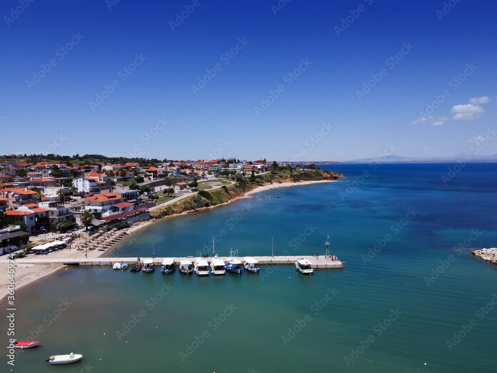 Chalkidiki, Greece coastal village landscape drone shot. Aerial day view of Nea Fokea seafront at Kassandra peninsula with low rise buildings by a clean tranquil blue sea, with moored fishing boats.