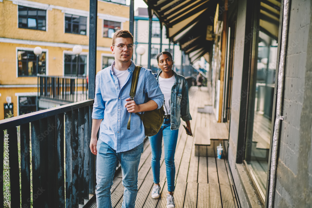 Young multiracial male and female international students 20s dressed in jeans trendy outfit walking outside of college building, diverse hipster teenagers in casual denim wear with backpacks