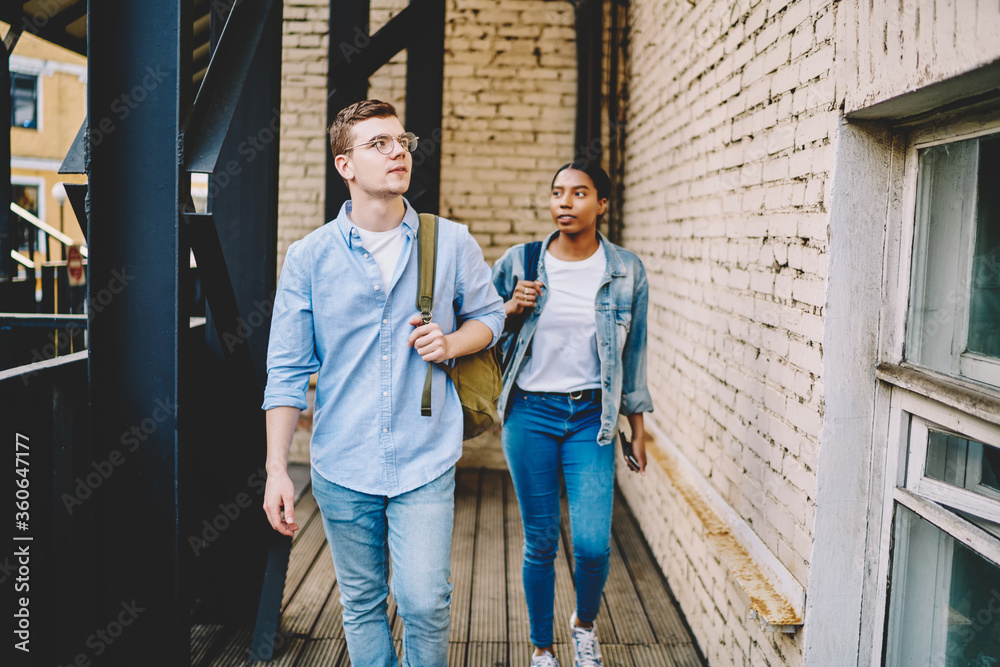 Obraz premium Young multiracial male and female students dressed in jeans trendy outfit walking in college building, teenagers in casual wear strolling with backpacks, hipster couple in stylish youth clothing