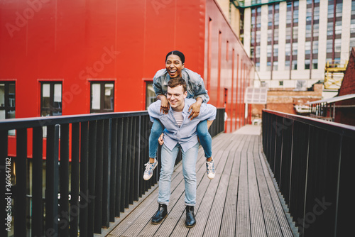 Happy African American hipster female laughing sitting at boyfriend's shoulder having fun outdoors, young couple in love spending free time together man give woman piggyback ride on street during date