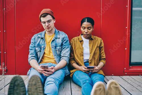 Multiracial couple resting together ignore communication concentrated on modern gadgets addiction and networking, dark skinned young woman jealous boyfriend looking at his smartphone sitting next