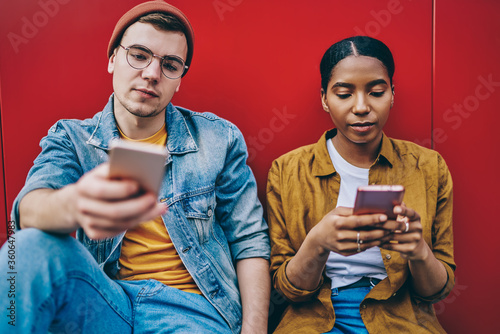 Trendy dressed multiracial hipster girl and guy sitting together ignore real communication prefere to chatting in social networks via modern smartphones and 4G internet connection, addiction to phone photo