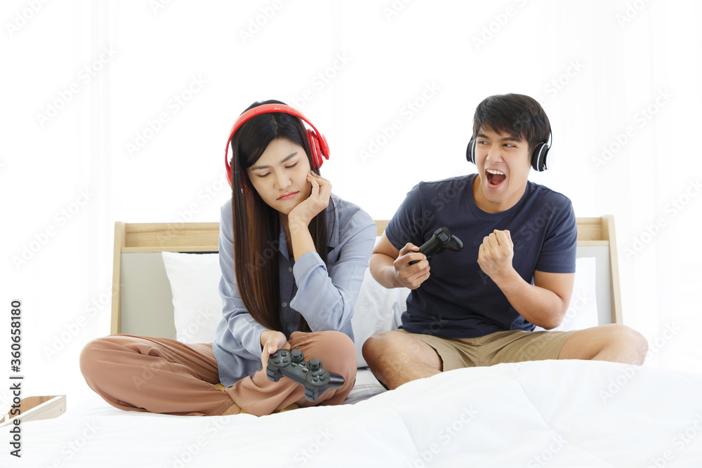 Young Asian Lovely Couple Playing Video Game console on bed in Bedroom. Spends time together. White Background. Male and Female. Bored Girl Friend.