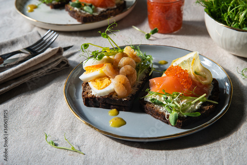 Smorrebrod dinner. Traditional Danish open rye bread sandwiches with smoked salmon and caviar and shrimps and egg, both with microgreens served on ceramic plates on linen tablecloth.
