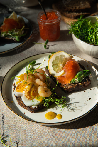 Smorrebrod dinner. Traditional Danish open rye bread sandwiches with smoked salmon and caviar and shrimps and egg, both with microgreens served on ceramic plates on linen tablecloth.