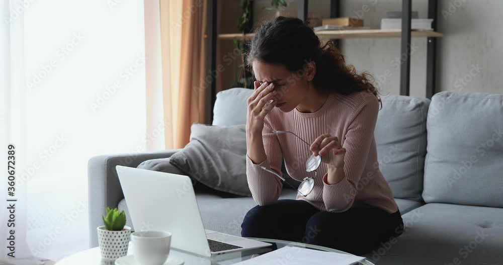 Overwhelmed frustrated young woman feeling tired due to computer overuse, suffering from blurred eyesight, massaging temples and irritated eyes. Stressed overworked female freelancer having migraine.