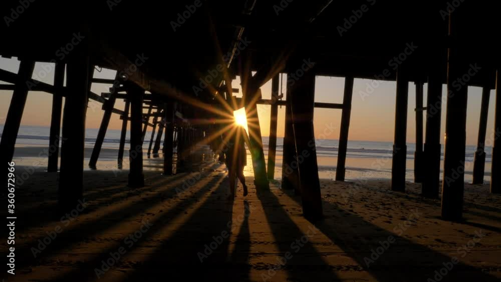 Woman silhouette walking under pier at Scenic orange pink Sunset with epic rays of light and sun flare wooden pier in San Clementa California Woman silhouette walking under pier at Scenic orange pink Sunset with epic rays of light and sun flare wooden pier in San Clementa California