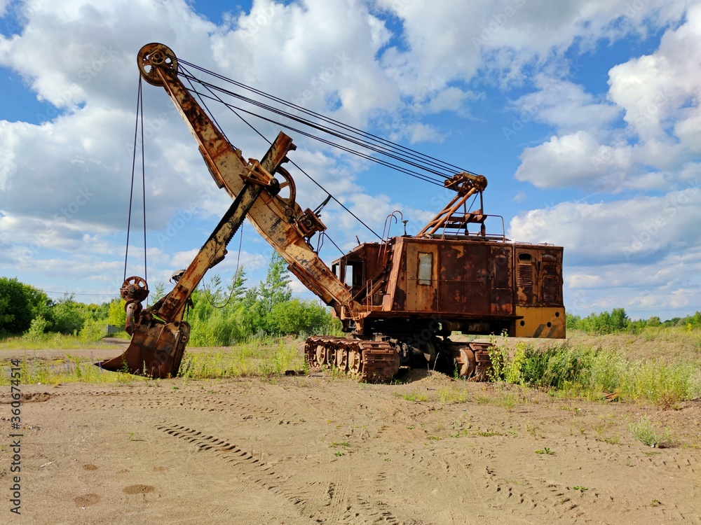 old career excavator with peeling paint stands on a sandy platform against the background of green grass and blue sky with clouds on a sunny day