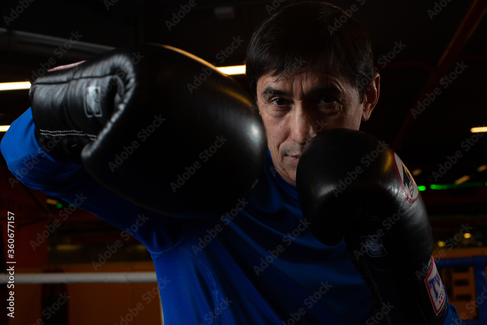 A man with black middle-aged hair in a blue rashgard and boxing gloves stands in a 'ready-to-attack' pose posing for the camera. Boxing club concept
