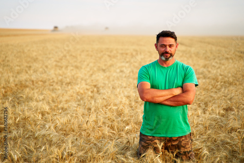 Happy farmer proudly standing in wheat field with arms crossed on chest. Agronomist wearing corporate uniform, looking at camera on farmland. Rich harvest of cultivated cereal crop. Harvesting season.