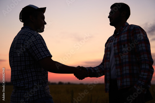 Farmer and agronomist silhouettes shaking hands standing in a wheat field after agreement in dusk. Agriculture business contract concept. Combine harvester driver and rancher handshake. Negotiations.