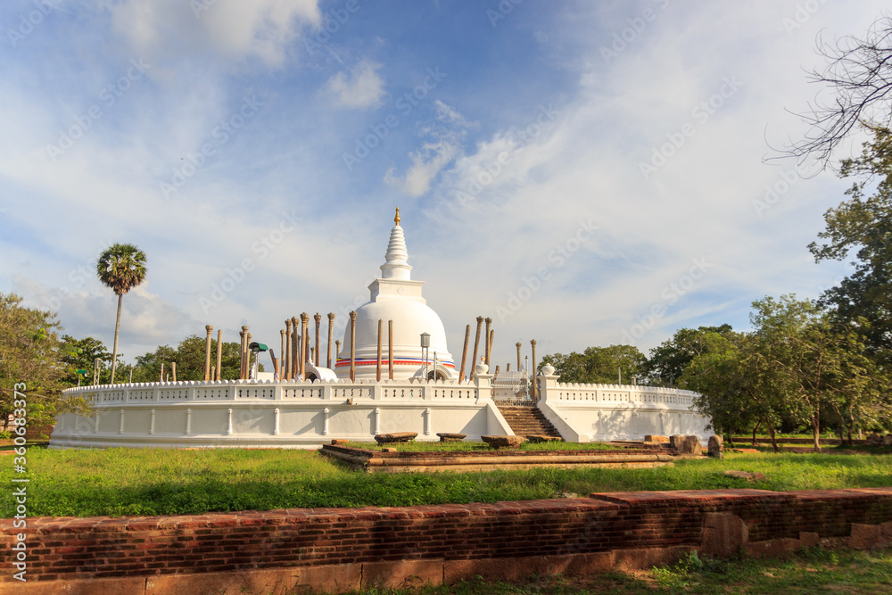 Fototapeta premium Panoramic view of restored Thuparamaya Dagoba with white bell shaped Stupa, altars, tilted vatadage pillars and Buddhist flags, Anuradhapura, Sri Lanka. Beautiful cloudy sky landscape.